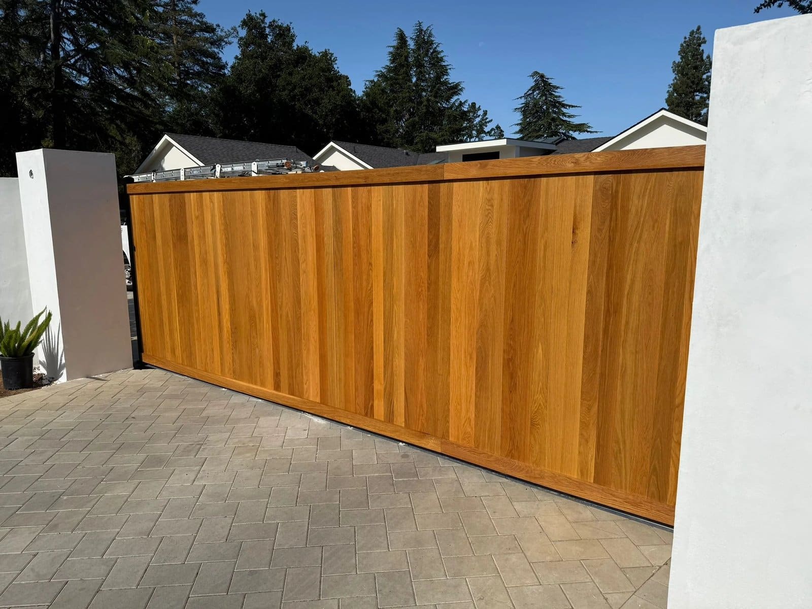 Custom wood sliding driveway gate with vertical cedar-tone boards between white stucco pillars on a gray paver driveway